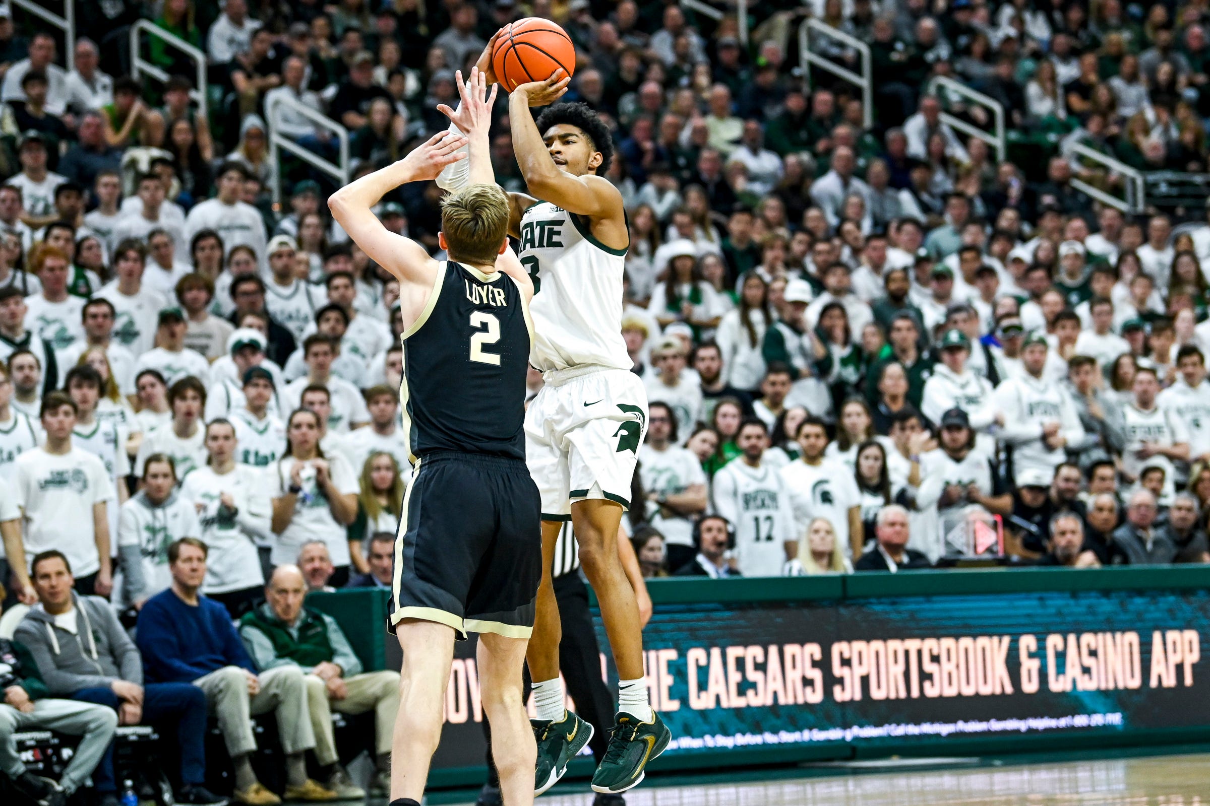 Michigan State's Jaden Akins makes a 3-pointer against Purdue's Fletcher Loyer. An advertisement for Caesars Underscoreg and Casino appears at the scorers table in the background.